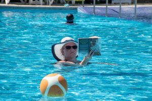 Lady in a pool with sunglasses and wide-brimmed hat, focused on reading.