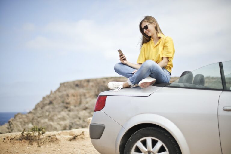 A happy girl sitting in the back of her convertible, browsing the internet with a smile.