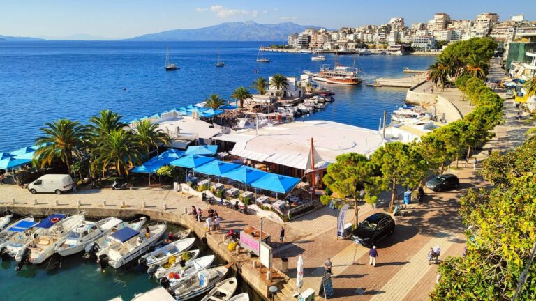 Harbor promenade with blue umbrellas, boats docked along a marina, and a hillside city in the distance.