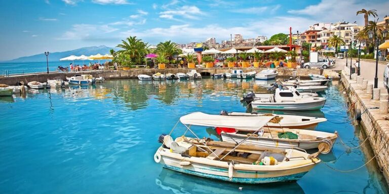 Boats docked in a calm turquoise harbor with a palm‑lined promenade and colorful buildings in the background.