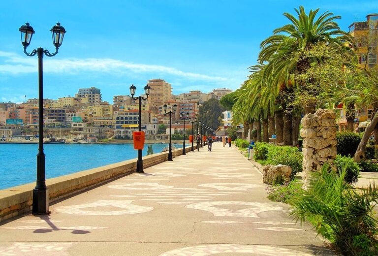 Seafront promenade with palm trees, vintage lampposts, and pastel buildings across the harbor. 細節: sunny day by the water.