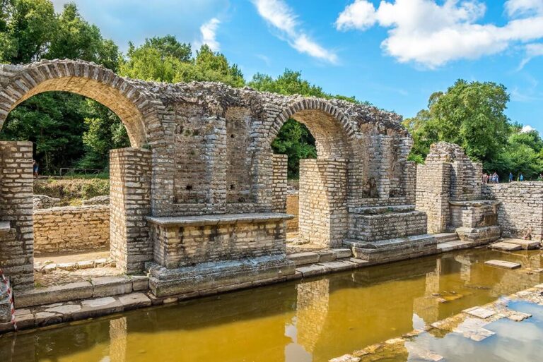 Ancient stone arches spanning a reflective pool, with green trees and a bright blue sky in the background