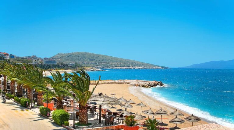Sunny beach with palm trees, wooden seating, and straw umbrellas along a sandy shore by bright blue water.