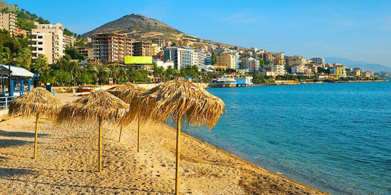 Beach with straw umbrellas on sandy shore, calm blue sea, and a colorful town along the coastline with hills in the background.